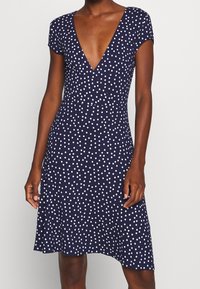 Kvinna i en marinblå klänning med vita prickar, korta ärmar och djup V-ringning, som står mot en enkel ljus bakgrund.
