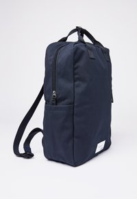 Laikku tummansininen kangaskassi, jossa on suorakulmainen muoto, vetoketjutuki, säädettävät hihnat ja sivutasku. Alhaalla on valkoinen etiketti.