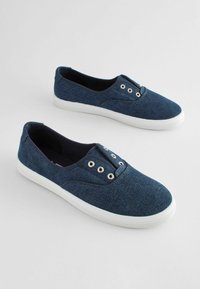 Mørkeblå canvas slip-on sko med hvide gummisåler, med metaløjer til snørebånd og en struktureret stofoverflade.