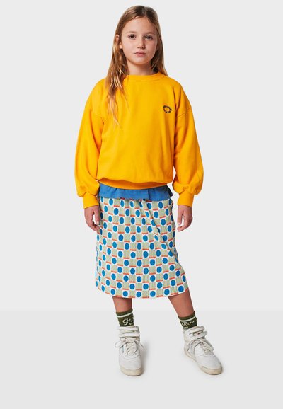 Gelber übergroßer Pullover, kombiniert mit einem blauen Oberteil, zu einem gemusterten Rock in Blau, Orange und Weiß. Weiße Sneakers und grüne Socken.