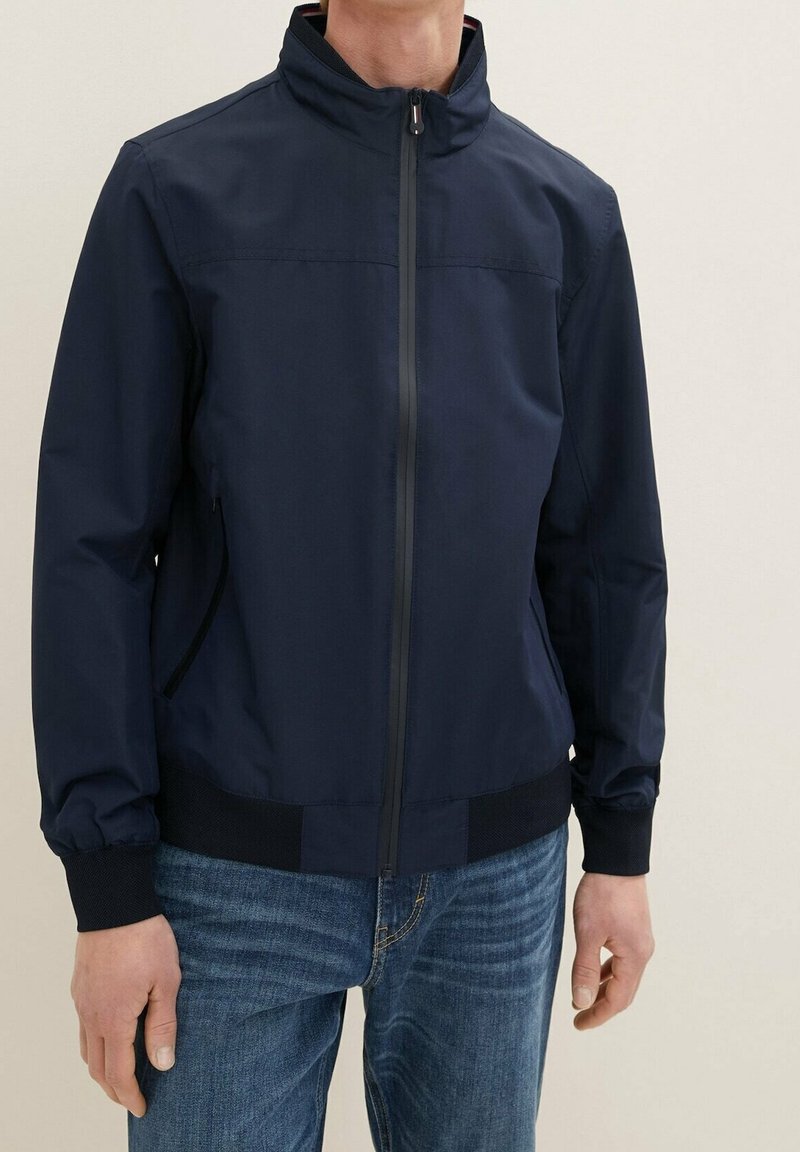 Homme portant une veste zippée bleu marine avec poignets et ourlet côtelés, associée à un jean bleu sur un fond uni.