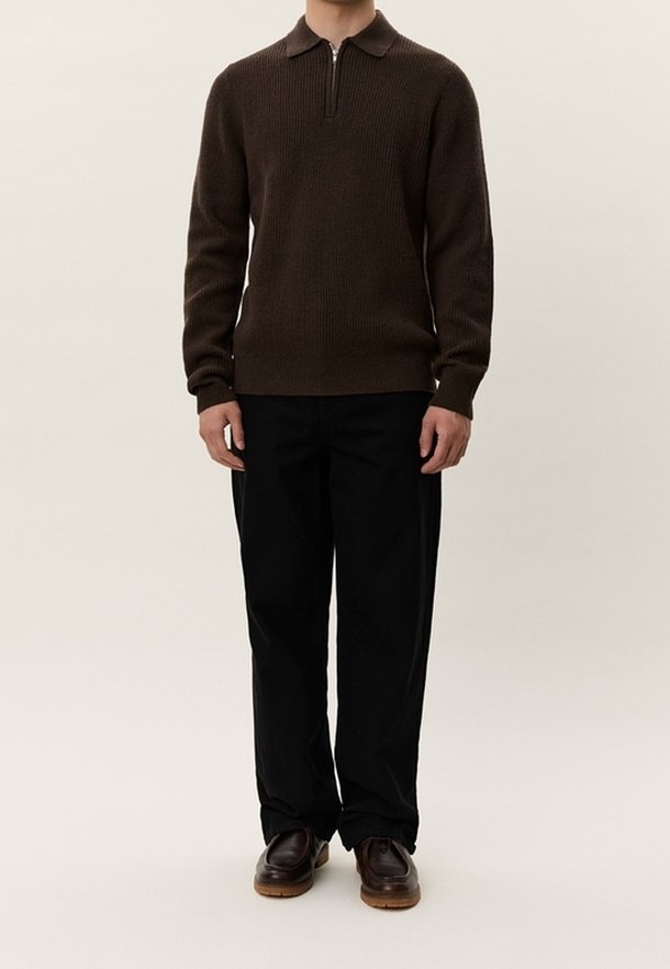 Homme portant un pull en maille marron foncé avec col et fermeture éclair, pantalon droit noir et chaussures en cuir marron foncé, debout devant un fond uni.