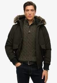 Veste à capuche noire avec bordure en fausse fourrure. Le modèle porte un pull en maille torsadée verte en dessous, associé à un jean bleu foncé et une ceinture noire.