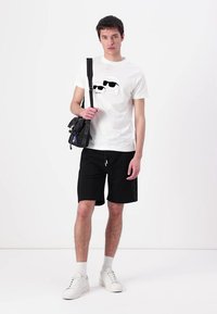 T-shirt bianca in cotone con stampa grafica nera, pantaloni corti neri e sneaker in pelle bianca. Il modello tiene in mano una piccola borsa a tracolla nera.