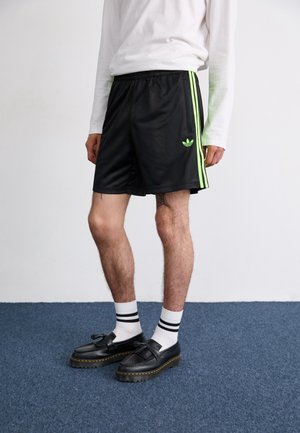 Personne portant un short de sport noir avec des rayures vert néon, une chemise blanche à manches longues, des chaussettes blanches avec des rayures noires et des chaussures en cuir noires.