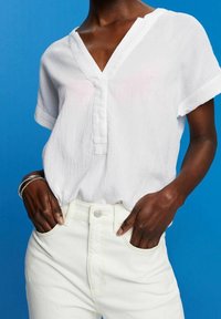 Weiße kurzärmlige Bluse mit V-Ausschnitt, aus strukturiertem Stoff mit Knopfdetails, kombiniert mit hellen hochgeschnittenen Jeans.