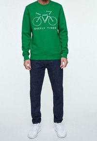 Osoba v zelené mikině se bílým grafikou bicyklu a textem "WHEELY TYRED", tmavých džínech a bílých teniskách, stojící čelem.