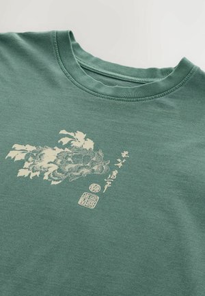 Camiseta de algodón verde con cuello redondo, que presenta un estampado floral crema y caracteres chinos en el frente. Textura suave, diseño casual.