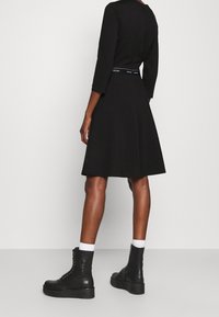 Černé šaty s tříčtvrtečními rukávy, vytaženým pasem s etikou Calvin Klein, rozšířenou sukní a v kombinaci s masivními černými šněrovacími botami.