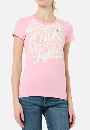 Frau trägt ein eng anliegendes pinkes T-Shirt mit weißem grafischem Schriftzug "BDK Fighter" und blaue Jeans vor einem einfarbigen weißen Hintergrund.