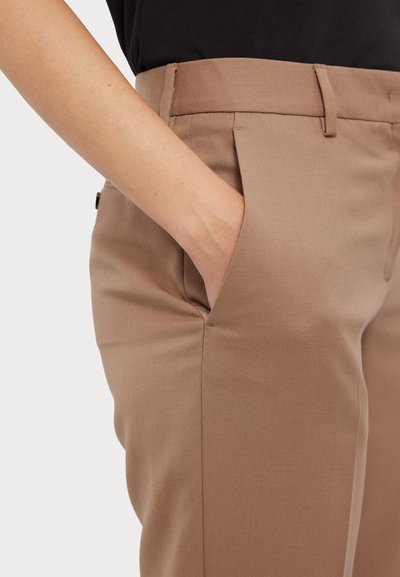 Bruine op maat gemaakte broek met een soepele textuur, voorzakken en riemlussen, weergegeven van taille tot hand in de zak.