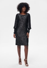 Czarna sukienka cekinowa o luźnym kroju, z długimi bufkami na rękawach, zaokrąglonym dekoltem i długością do kolan. Stylizowana z cielistymi sandałami na paski.