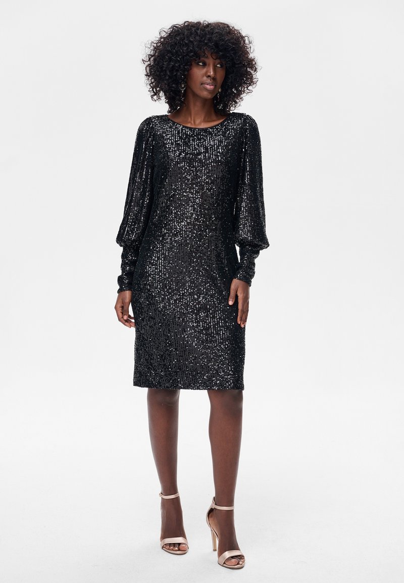 Czarna sukienka cekinowa o luźnym kroju, z długimi bufkami na rękawach, zaokrąglonym dekoltem i długością do kolan. Stylizowana z cielistymi sandałami na paski.