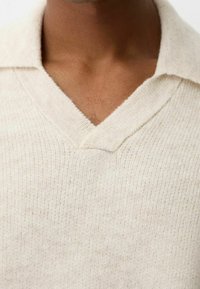 Gros plan sur une personne portant un pull en maille de couleur crème avec un col en V et un tissu doux et texturé.