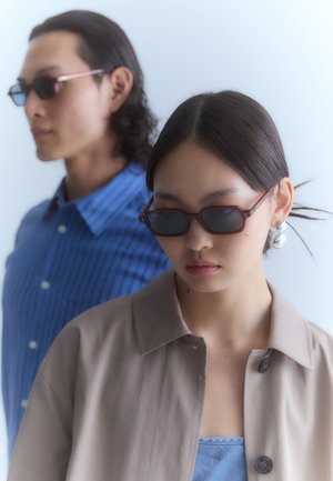 Brown rectangular sunglasses with dark lenses. One person wears a beige collared jacket; the other wears a blue striped shirt.