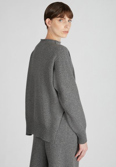 Grauer Strickpullover mit hohem Kragen, überschnittenen Schultern und geripptem Saum. Das Material wirkt weich mit einer strukturierten Oberfläche.