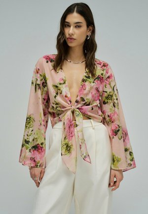Femme aux cheveux bruns ondulés portant un haut transparent à manches longues rose à motifs floraux noué à l'avant et un pantalon blanc taille haute, sur un fond neutre.