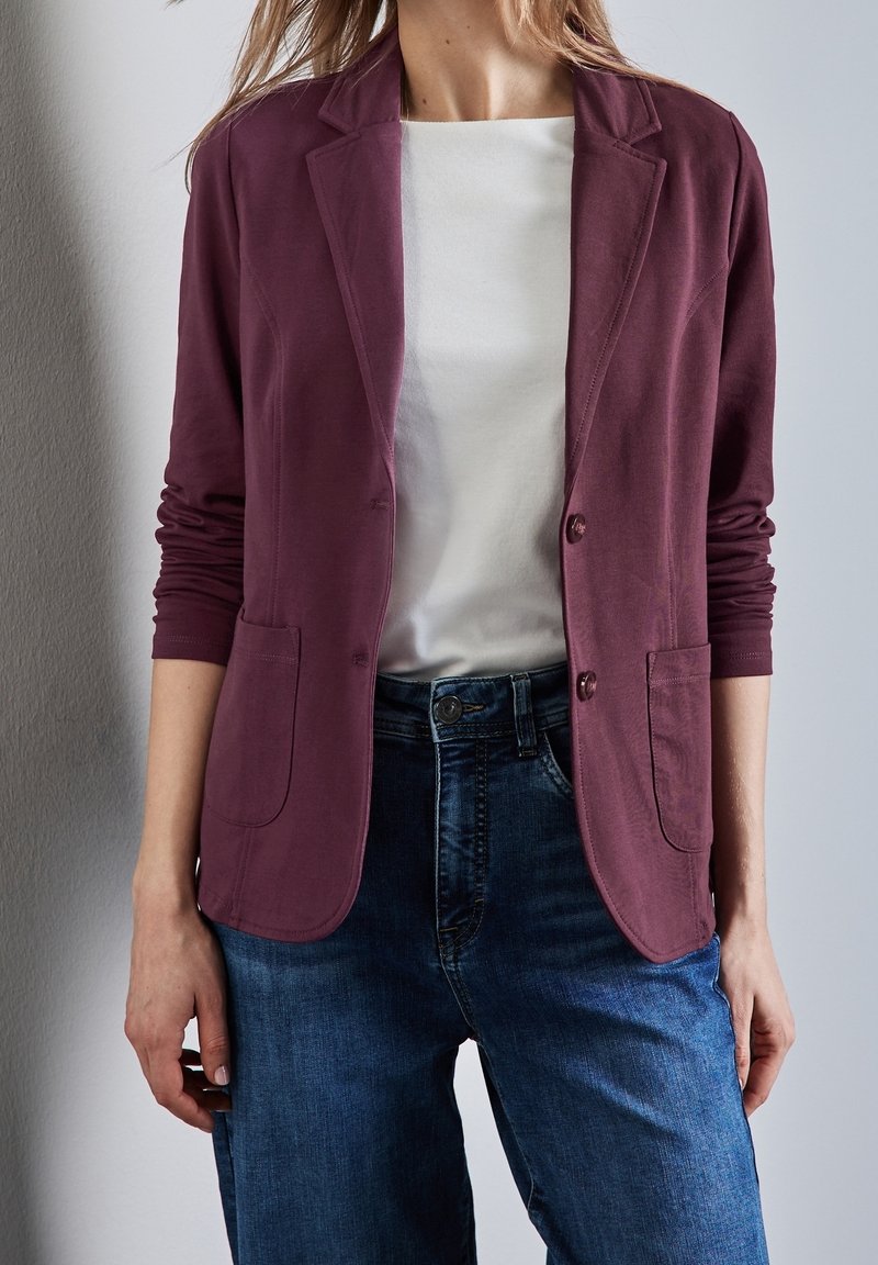 Donna che indossa un blazer prugna con maniche arrotolate, una maglietta bianca e jeans blu, in piedi contro un muro grigio chiaro.
