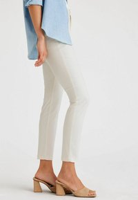 Pantaloni bianchi aderenti con una silhouette slim e cuciture visibili, abbinati a sandali con tacco blocco color cenere. La parte superiore è una camicia azzurra chiara.