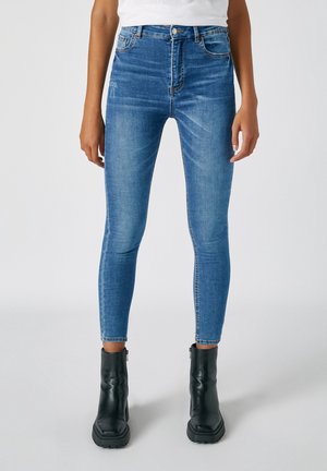 Personne portant un jean bleu slim et des bottines noires, debout devant un fond clair uni.