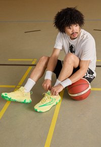 Basketbalové boty žluté barvy s zelenými akcenty, vyrobené z prodyšného materiálu a s texturovanou podrážkou. Nosené s bílými sportovními ponožkami a míčem.