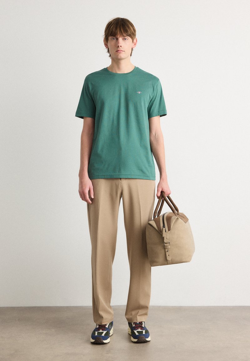 Grünes Baumwoll-T-Shirt, beige Hose und braune strukturierte Tasche. Sneaker mit blauen, weißen und roten Akzenten. Schlichtes, lockeres Design.