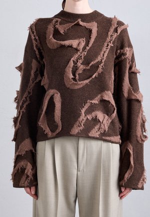 Pull en maille marron avec des motifs de volants irréguliers en relief, porté avec un pantalon large plissé beige.