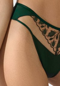String vert avec des panneaux en mesh transparent ornés de broderies florales noires. Texture de tissu lisse avec des bords galbés et un léger éclat.