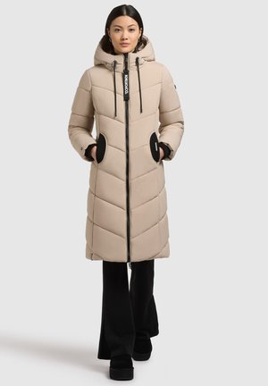Femme portant un manteau doudoune beige long avec capuche, mains dans des poches à bord noir, pantalon noir et chaussures à plateforme noires, se tenant sur un fond blanc.