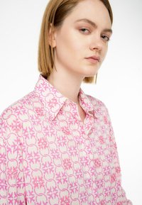 Camicia button-up rosa chiaro con un motivo floreale geometrico in rosa scuro, completa di decorazioni floreali testurizzate sulle spalle.