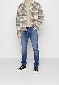 Rutigt ullskjortjacka i krämfärgad och brun, beige t-shirt, blå denimjeans och svarta sneakers med vita sulor och snören.