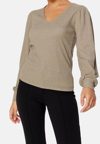 Donna che indossa una blusa beige a maniche lunghe con scollatura a V e texture, abbinata a pantaloni neri aderenti, mostrata dalla nuca ai fianchi.