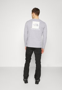 Langärmliges graues T-Shirt mit einem aufgedruckten weißen Logo und Text auf dem Rücken, kombiniert mit schwarzen Hosen und schwarzem Schuhwerk. Glatte Textur.