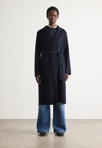 Mulher vestindo um casaco azul-marinho cinto sobre um suéter escuro e uma camisa de gola, combinado com jeans de perna larga azuis e sapatos pretos, em pé sobre um chão simples.