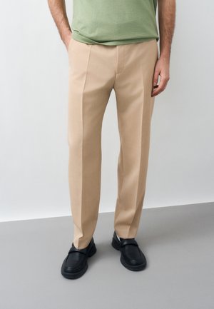 Muž nosiaci béžové šité nohavice, zelené tričko a čierne slip-on topánky stojaci pred jednofarebným pozadím.