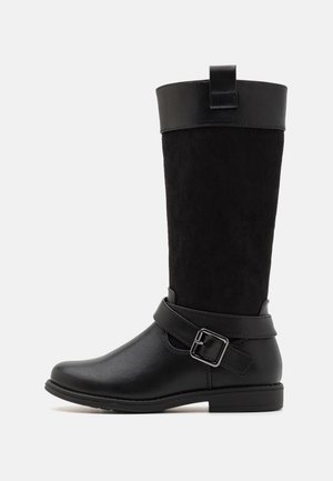 Botte noire mi-mollet avec pied en cuir lisse, tige en suède, une sangle avec boucle en métal autour de la cheville, et une languette de tirage en haut.