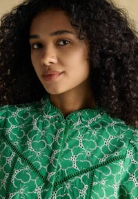 Blusa verde ricamata caratterizzata da intricati motivi floreali, scollatura a punta e accenti bianchi a contrasto. Tessuto leggero e con texture.