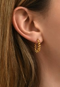 Boucle d'oreille en or tordu avec un design en spirale texturé, mise en valeur sur un teint de peau claire et partiellement obscurcie par les cheveux.