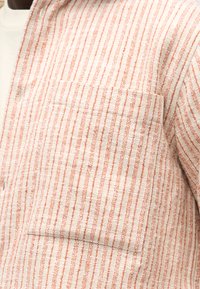 Μπεζ και πορτοκαλί πουκάμισο με κάθετες ρίγες και θήκη στο στήθος πάνω από λευκό εσώρουχο, που δείχνει την υφή του υφάσματος και το κούμπωμα με κουμπιά.