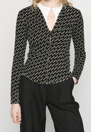 Frau trägt eine schwarze Reißverschlussjacke mit wiederholtem "PINKO"-Logomuster über einem weißen Top mit Schlüssellochausschnitt, kombiniert mit schwarzen Hosen.