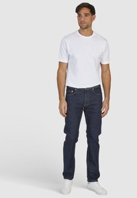 Weißes Kurzarm-T-Shirt kombiniert mit dunkelblauen Jeans und weißen Turnschuhen. Die Jeans verfügen über traditionelle Taschen und dezente Steppdetails.