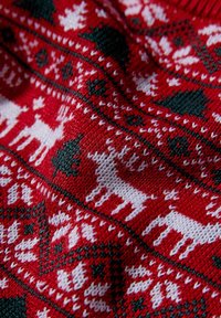 Röd stickad tyg med vita renar och gröna träd i ett upprepande mönster, med en strukturerad yta och kontrasterande mörka detaljer.