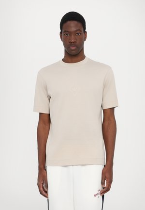T-shirt in cotone beige con maniche corte, scollo rotondo e un discreto logo a forma di diamante sul petto. Tessuto morbido, vestibilità comoda.