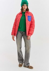 Žena si oblékla zelenou čepici, zelený svetr, korálově prošívanou bundu s modrým kapsou, šedé džíny a boty s leopardím vzorem, stojící na bílém pozadí.