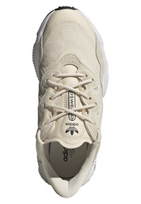 Beige sneaker with textured mesh and smooth leather accents. Features a round toe, flat laces, and a white cushioned sole. Logo on tongue.
