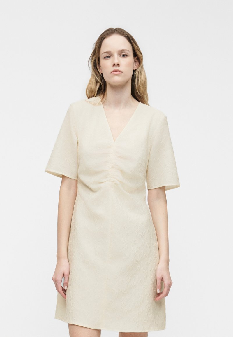 Femme debout devant un fond uni, portant une robe à manches courtes de couleur crème avec un col en V et un motif texturé subtil.