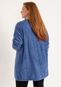 Fuzzy, mid-length cardigan in soft blue fabric. Features wide sleeves, an open silhouette, and a textured surface with a relaxed fit.