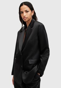 Blazer in raso nero con colletto a scialle, chiusura con un bottone e due tasche frontali, che mostra una consistenza liscia e lucida.