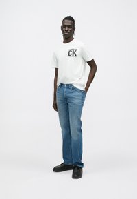 Biała bawełniana koszulka z czarnym napisem i logo "CALVIN KLEIN", w połączeniu z niebieskimi jeansami i czarnymi trampkami, stojąca na jednolitym tle.