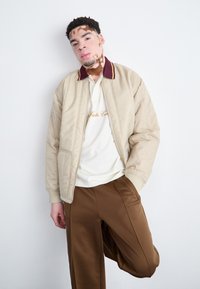 Beige Bomberjacke mit bordeauxfarbenem Kragen, mit Reißverschluss vorne; hellfarbiges Polo mit goldenen Stickereien; braune Hose mit Seitentaschen.